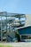 A partially collapsed metal structure containing several boats stacked on different levels, with some sections of the building visibly damaged. The scene includes a blue building with a window that has 'Apparel Accessories' written on it. There is a palm tree next to the building, and the ground appears to be covered with debris.