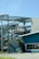 A partially collapsed metal structure containing several boats stacked on different levels, with some sections of the building visibly damaged. The scene includes a blue building with a window that has 'Apparel Accessories' written on it. There is a palm tree next to the building, and the ground appears to be covered with debris.