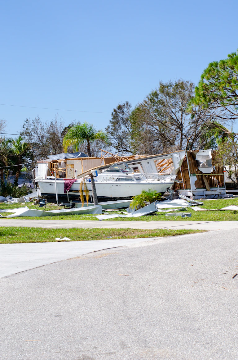 a boat that is sitting in the grass against a mobile home destroyed in a hurricane