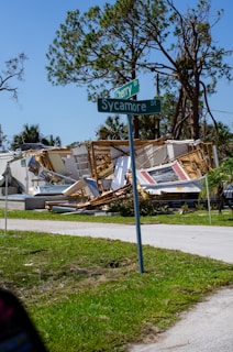 A collapsed house with debris scattered around, including sections of walls and roof. In the foreground, a street sign shows the intersection of Cherry Drive and Sycamore Street. Large trees with some broken branches are in the background under a clear blue sky.