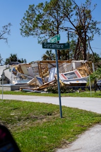 A collapsed house with debris scattered around, including sections of walls and roof. In the foreground, a street sign shows the intersection of Cherry Drive and Sycamore Street. Large trees with some broken branches are in the background under a clear blue sky.