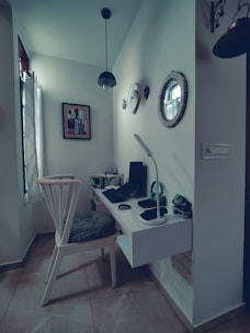 A cozy corner workspace featuring a minimalistic floating desk with a modern chair. The desk is equipped with a computer, desk lamp, and stationary items. Wall decorations include a round mirror, a clock, and a piece of framed art. Pendant lighting hangs from the ceiling, complementing the natural light streaming through a window with sheer curtains.