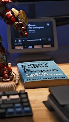 A desktop setup featuring a book titled 'Everything is F*cked: A Book About Hope' by Mark Manson. Beside the book is a figurine of a red and gold armored hand, possibly from a superhero. In the background, a smart display shows a music application playing Metallica's 'The Unforgiven'. Additional objects include part of a computer keyboard and a coiled cable.