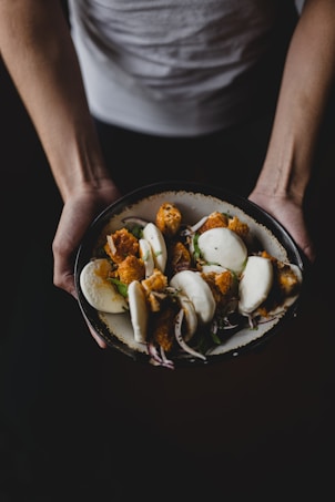 A person is holding a dark bowl filled with white steamed buns stuffed with crispy fried fillings, garnished with fresh herbs and sliced onions. The scene is captured with dramatic lighting, highlighting the textures and colors of the food.