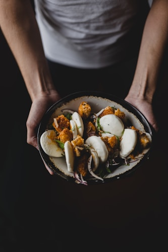 A person is holding a dark bowl filled with white steamed buns stuffed with crispy fried fillings, garnished with fresh herbs and sliced onions. The scene is captured with dramatic lighting, highlighting the textures and colors of the food.