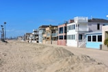 A row of beach houses with varied architectural styles lines a sandy beachfront under a clear blue sky. The houses feature multiple stories, large windows, balconies, and pastel colors. Tall palm trees are visible to the left, adding to the coastal setting.