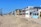 A row of beach houses with varied architectural styles lines a sandy beachfront under a clear blue sky. The houses feature multiple stories, large windows, balconies, and pastel colors. Tall palm trees are visible to the left, adding to the coastal setting.