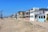 A row of beach houses with varied architectural styles lines a sandy beachfront under a clear blue sky. The houses feature multiple stories, large windows, balconies, and pastel colors. Tall palm trees are visible to the left, adding to the coastal setting.