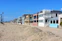 A row of beach houses with varied architectural styles lines a sandy beachfront under a clear blue sky. The houses feature multiple stories, large windows, balconies, and pastel colors. Tall palm trees are visible to the left, adding to the coastal setting.