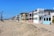A row of beach houses with varied architectural styles lines a sandy beachfront under a clear blue sky. The houses feature multiple stories, large windows, balconies, and pastel colors. Tall palm trees are visible to the left, adding to the coastal setting.