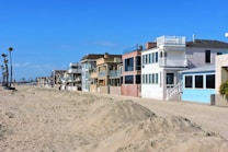 A row of beach houses with varied architectural styles lines a sandy beachfront under a clear blue sky. The houses feature multiple stories, large windows, balconies, and pastel colors. Tall palm trees are visible to the left, adding to the coastal setting.