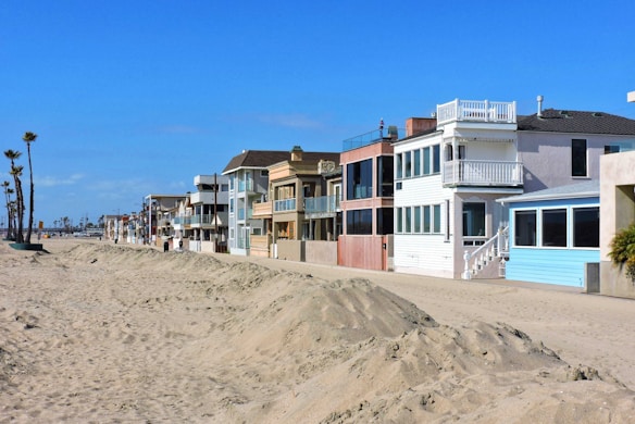 A row of beach houses with varied architectural styles lines a sandy beachfront under a clear blue sky. The houses feature multiple stories, large windows, balconies, and pastel colors. Tall palm trees are visible to the left, adding to the coastal setting.