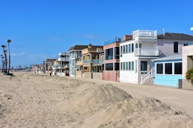 A row of beach houses with varied architectural styles lines a sandy beachfront under a clear blue sky. The houses feature multiple stories, large windows, balconies, and pastel colors. Tall palm trees are visible to the left, adding to the coastal setting.