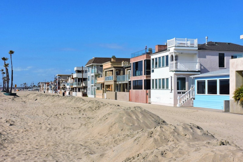 A row of beach houses with varied architectural styles lines a sandy beachfront under a clear blue sky. The houses feature multiple stories, large windows, balconies, and pastel colors. Tall palm trees are visible to the left, adding to the coastal setting.