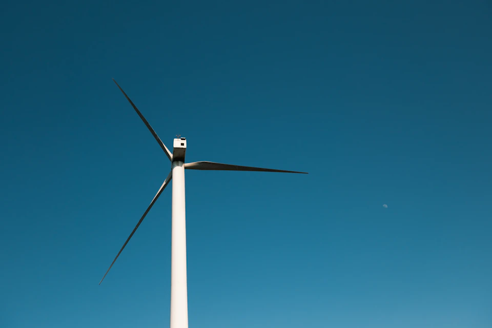 The image depicts a large white wind turbine standing tall against a clear blue sky. The turbine has three blades, each pointing in different directions, suggesting it is actively harnessing the power of the wind to generate electricity. In the top right corner of the photo, there is a small moon visible, adding an element of natural beauty to the scene.

The image does not contain any discernible text or human-made objects, and the focus remains solely on the wind turbine and its surroundings.