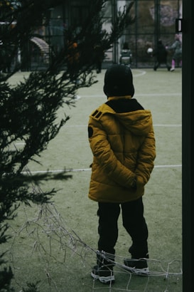 A child in a yellow jacket, black pants, and hat stands on an outdoor sports court with hands behind their back. The scene is framed by dark green foliage, with a few people and a soccer ball visible in the background, giving a sense of a recreational environment.