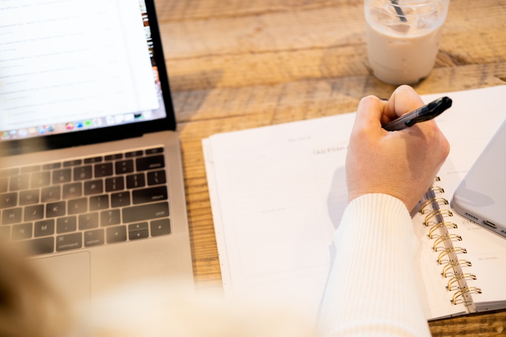 a person writing on a notebook next to a laptop