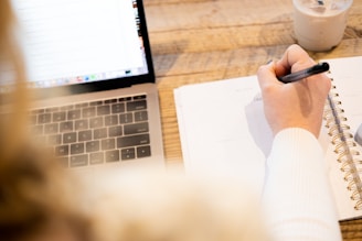 a person writing on a notebook next to a laptop