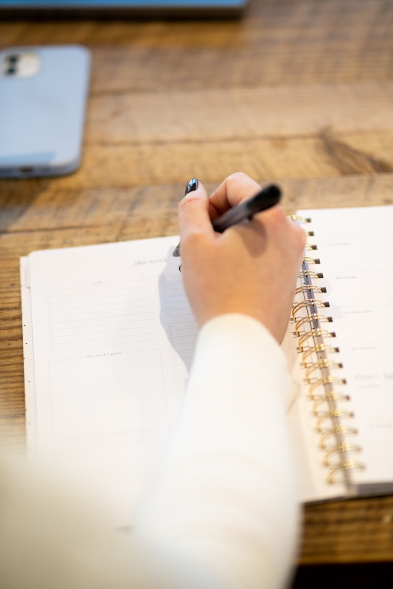 A hand writing in a planner beside a phone on a wooden table