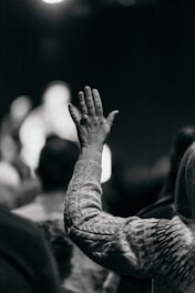 A monochrome image featuring a person's hand raised in a crowd, suggesting engagement or participation. The focus is on the hand, which is adorned with a ring, while the rest of the image is blurred, indicating a setting with multiple people.