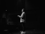 A person stands on a stage in a dimly lit environment, facing the audience. They appear to be speaking, with one hand slightly raised. The setting suggests a presentation or performance, with minimal lighting highlighting the speaker.