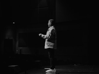 A person stands on a stage in a dimly lit environment, facing the audience. They appear to be speaking, with one hand slightly raised. The setting suggests a presentation or performance, with minimal lighting highlighting the speaker.