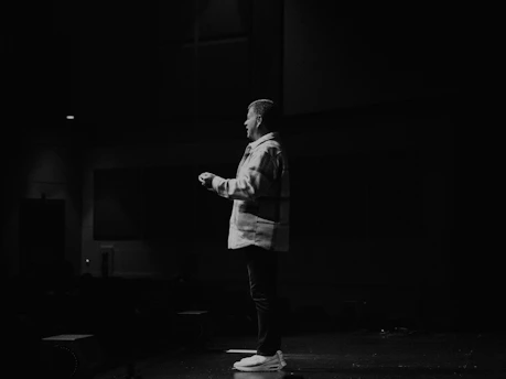 A dimly lit stage with a speaker addressing an attentive audience in a moody atmosphere.