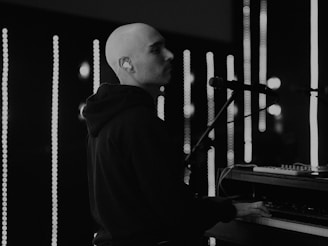 A person is playing a keyboard or piano in a dimly lit setting with vertical light bars in the background. The individual is wearing a hoodie and appears focused on the music. The scene has a monochrome tone, emphasizing contrasts between the dark clothing and the bright lights.