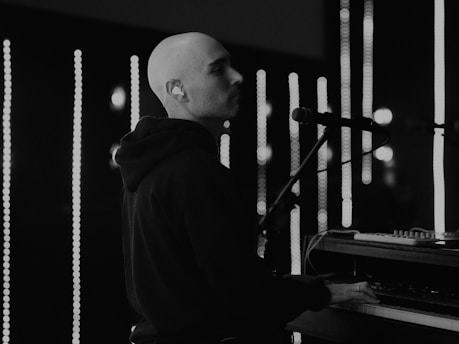 A person is playing a keyboard or piano in a dimly lit setting with vertical light bars in the background. The individual is wearing a hoodie and appears focused on the music. The scene has a monochrome tone, emphasizing contrasts between the dark clothing and the bright lights.