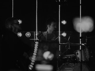 A monochromatic scene featuring two musicians performing. One is playing the drums, and the other appears to be playing a guitar or bass. Both are surrounded by bright lights in the background, which create a vibrant, yet dramatic setting. The focus on their instruments suggests a live performance or practice session.