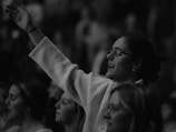 A group of people, primarily women, appears to be engaged in an emotional or spiritual moment. One woman with glasses in the foreground is raising her arm, possibly in a gesture of praise or worship.