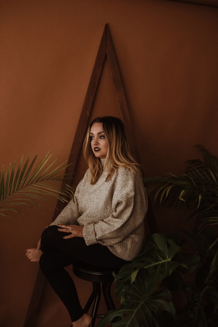 A person with long, wavy hair poses seated on a stool, surrounded by green foliage. The background features a warm brown hue with a triangular wooden decoration, creating a cozy and natural ambiance. The person wears a beige sweater and black pants, exuding a calm and introspective demeanor.