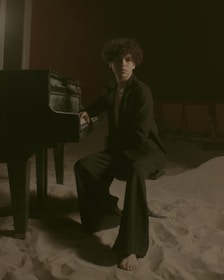 A person with curly hair sits barefoot at a piano in a dimly lit space covered with sand. The environment is unusual, resembling an indoor setting with theatrical seats in the background and a moody ambiance.