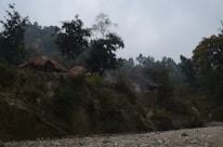 A rural landscape featuring a couple of thatched huts situated on a hill covered with trees and vegetation. The sky is overcast, lending a moody atmosphere to the scene. The foreground shows a rocky and pebbly riverbank or dry riverbed.