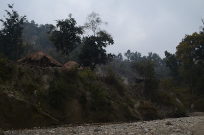 A rural landscape featuring a couple of thatched huts situated on a hill covered with trees and vegetation. The sky is overcast, lending a moody atmosphere to the scene. The foreground shows a rocky and pebbly riverbank or dry riverbed.