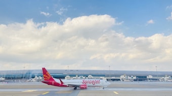 A SpiceJet airplane is positioned on the runway with a large modern terminal building in the background. The sky is mostly clear with a few scattered clouds. The airplane is white with a red tail that features a yellow design.