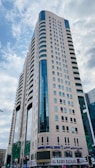 A tall modern skyscraper with a beige facade and blue-tinted windows dominates the scene. The building features several rows of windows and appears to have a commercial area on the ground floor, including a sign for 'AL RAMS BAZAAR'. The sky is partially cloudy.