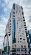 A tall modern skyscraper with a beige facade and blue-tinted windows dominates the scene. The building features several rows of windows and appears to have a commercial area on the ground floor, including a sign for 'AL RAMS BAZAAR'. The sky is partially cloudy.