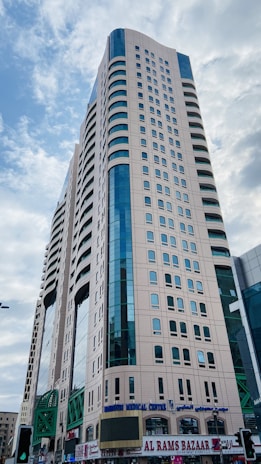 A tall modern skyscraper with a beige facade and blue-tinted windows dominates the scene. The building features several rows of windows and appears to have a commercial area on the ground floor, including a sign for 'AL RAMS BAZAAR'. The sky is partially cloudy.