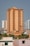 A tall, beige and brown high-rise building stands prominently in an urban cityscape. The structure is surrounded by other similar residential buildings and is accompanied by lush green palm trees at its base.