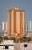 A tall, beige and brown high-rise building stands prominently in an urban cityscape. The structure is surrounded by other similar residential buildings and is accompanied by lush green palm trees at its base.