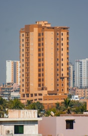A tall, beige and brown high-rise building stands prominently in an urban cityscape. The structure is surrounded by other similar residential buildings and is accompanied by lush green palm trees at its base.