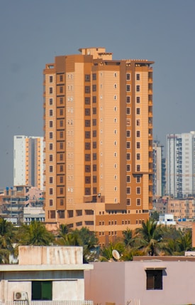 A tall, beige and brown high-rise building stands prominently in an urban cityscape. The structure is surrounded by other similar residential buildings and is accompanied by lush green palm trees at its base.