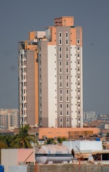 A tall, modern apartment building with a mix of beige, white, and light brown colors. It features numerous windows and is surrounded by smaller buildings and some palm trees. The sky is clear and there is a large water tank visible on one of the rooftops in the foreground.
