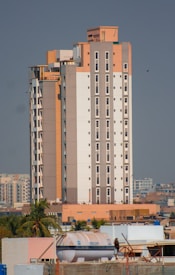 A tall, modern apartment building with a mix of beige, white, and light brown colors. It features numerous windows and is surrounded by smaller buildings and some palm trees. The sky is clear and there is a large water tank visible on one of the rooftops in the foreground.