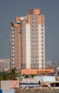 A tall, modern apartment building with a mix of beige, white, and light brown colors. It features numerous windows and is surrounded by smaller buildings and some palm trees. The sky is clear and there is a large water tank visible on one of the rooftops in the foreground.