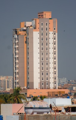 A tall, modern apartment building with a mix of beige, white, and light brown colors. It features numerous windows and is surrounded by smaller buildings and some palm trees. The sky is clear and there is a large water tank visible on one of the rooftops in the foreground.