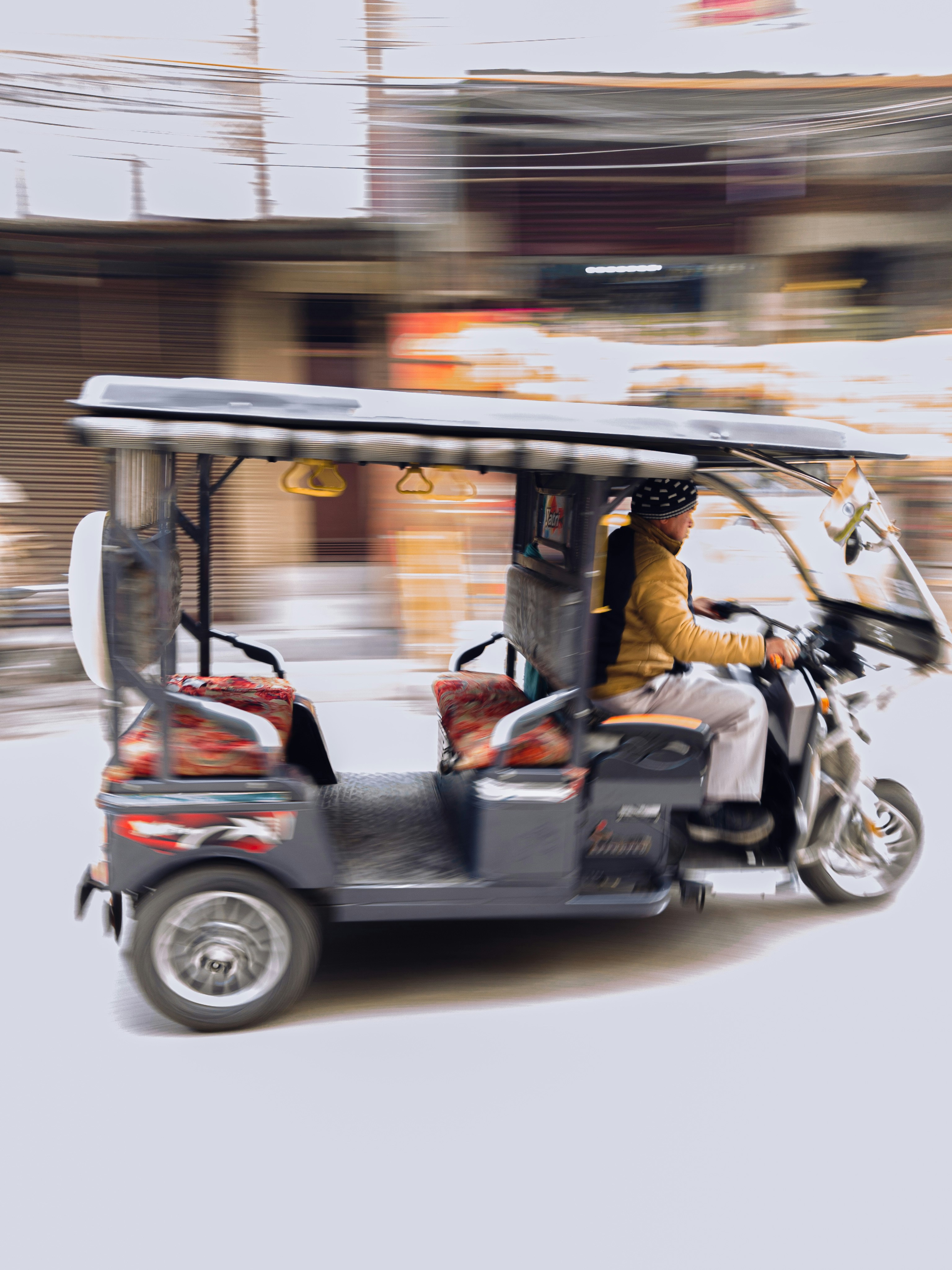 A motorized rickshaw is moving quickly down a street. The blurred background suggests motion and speed. The rickshaw is carrying a person who is bundled up in warm clothing, with decorative cushions on the passenger seats.