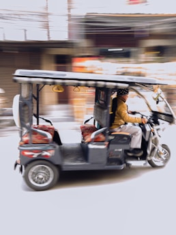 A motorized rickshaw is moving quickly down a street. The blurred background suggests motion and speed. The rickshaw is carrying a person who is bundled up in warm clothing, with decorative cushions on the passenger seats.