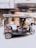 A motorized rickshaw is moving quickly down a street. The blurred background suggests motion and speed. The rickshaw is carrying a person who is bundled up in warm clothing, with decorative cushions on the passenger seats.